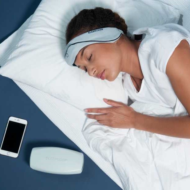 Woman lying in bed with a device covering her ears and forehead, beside her a white container and her smartphone